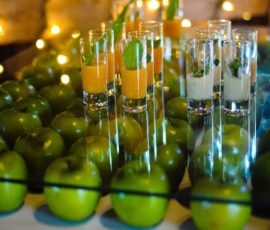 Rows of green apples surround glasses of orange and white drinks, garnished with mint, on a glass-topped table.