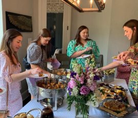 Four women serve themselves food from a buffet table with flowers and assorted dishes in a bright dining room.