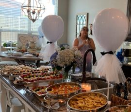 A kitchen island is covered with assorted buffet-style food trays; white balloons and flowers decorate the space.