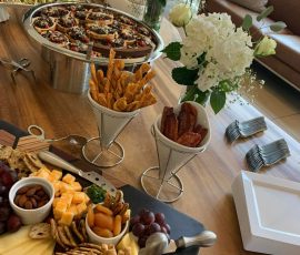 Cheese and charcuterie board with crackers, breadsticks, and flowers set on a wooden table in a modern living room.