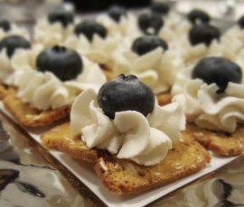 Screenshot Toasted crackers topped with swirls of whipped cream and a single blueberry on each, arranged on a reflective surface.