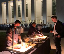 Two chefs serve food to a man at an outdoor buffet event in front of a large, well-lit building at night.