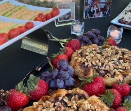 A table with fruit, cheese, and nut-topped pastries, with photos and candles, set outdoors under a tree.