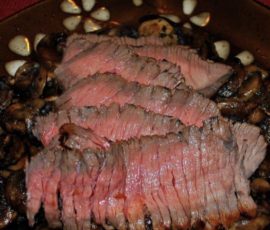 Sliced medium-rare roast beef served on a bed of sautéed mushrooms, garnished with garlic on a brown plate.