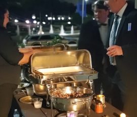 Three people in formal attire stand at an outdoor buffet table at night, serving food from chafing dishes.