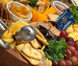 A wooden tray with assorted cheeses, crackers, fruit, dried fruit, and garnish, arranged for serving.