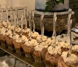 Mini dessert cups with whipped topping are arranged in front of a two-tiered white and blue cake with floral decor.