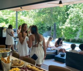 Women in white dresses gather inside and outdoors by a pool; a table is set with fruit, vegetables, and snacks.