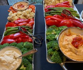 Two trays of assorted fresh vegetables with bowls of hummus and dip, set on a table by a window.