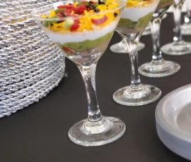 Several layered dip appetizers are served in martini glasses on a black table next to a stack of silver placemats.