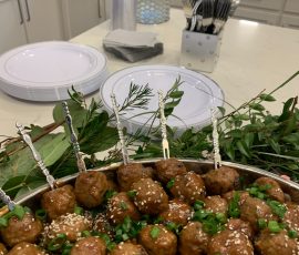 A platter of sesame meatballs garnished with green onions, with plates, utensils, and drinks in the background.