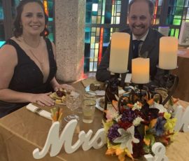 Two people in formal attire sit at a decorated table with candles and flowers in front of stained glass windows.
