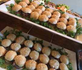 Two trays of mini burgers with sesame seed buns are arranged on lettuce, with assorted fruit visible in the background.