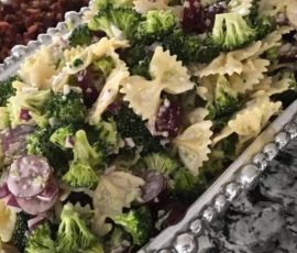 Bow tie pasta salad with broccoli, red onions, and dressing served on a silver platter on a granite countertop.