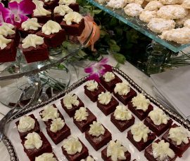 Plates of red velvet cake squares with white frosting and powdered sugar cookies displayed on a table with floral decor.