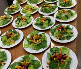 Rows of white plates with green salads topped with corn, black beans, and diced bell peppers on a wooden table.