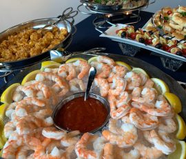 Chilled shrimp with cocktail sauce and lemon wedges, surrounded by assorted hot appetizers on serving trays.